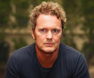 A man with curly hair in a serious expression wearing a black shirt, with a blurred outdoor background.