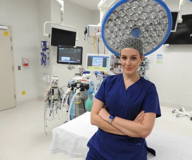 A nurse in blue scrubs stands confidently in an operating room with medical equipment.
