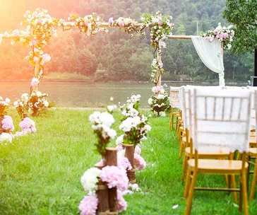 Outdoor wedding setup with floral arch and chairs by a river, under a soft sunlight glow.