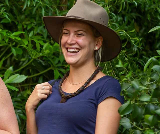 Woman in a hat smiling, surrounded by greenery.