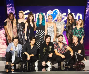 A group of 14 individuals smiling, posing together on stage in front of a Eurovision logo backdrop.
