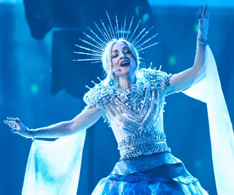Performer in an elaborate silver costume with a crown-like headpiece sings passionately on stage under blue lighting.