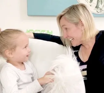 A woman and a young girl share a joyful conversation on a white sofa, smiling at each other warmly.