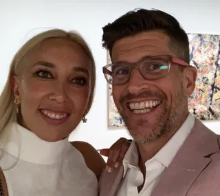 A couple smiles together while taking a selfie, with art visible in the background.