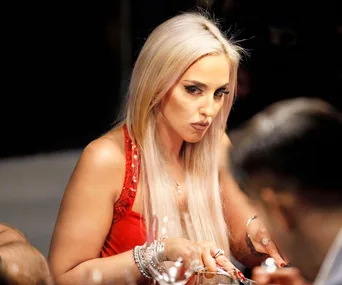A woman with long blonde hair, wearing a red dress, is sitting at a table cutting food, looking serious.
