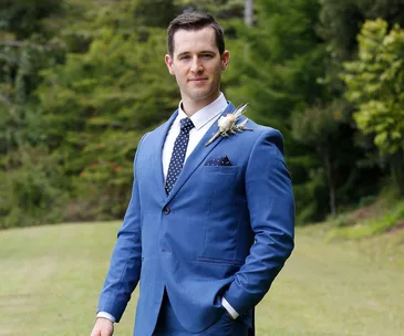 Man in a blue suit and polka dot tie stands outdoors with greenery in the background.
