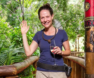 Smiling woman in a blue shirt holding a glass of champagne, waving in a lush outdoor setting.