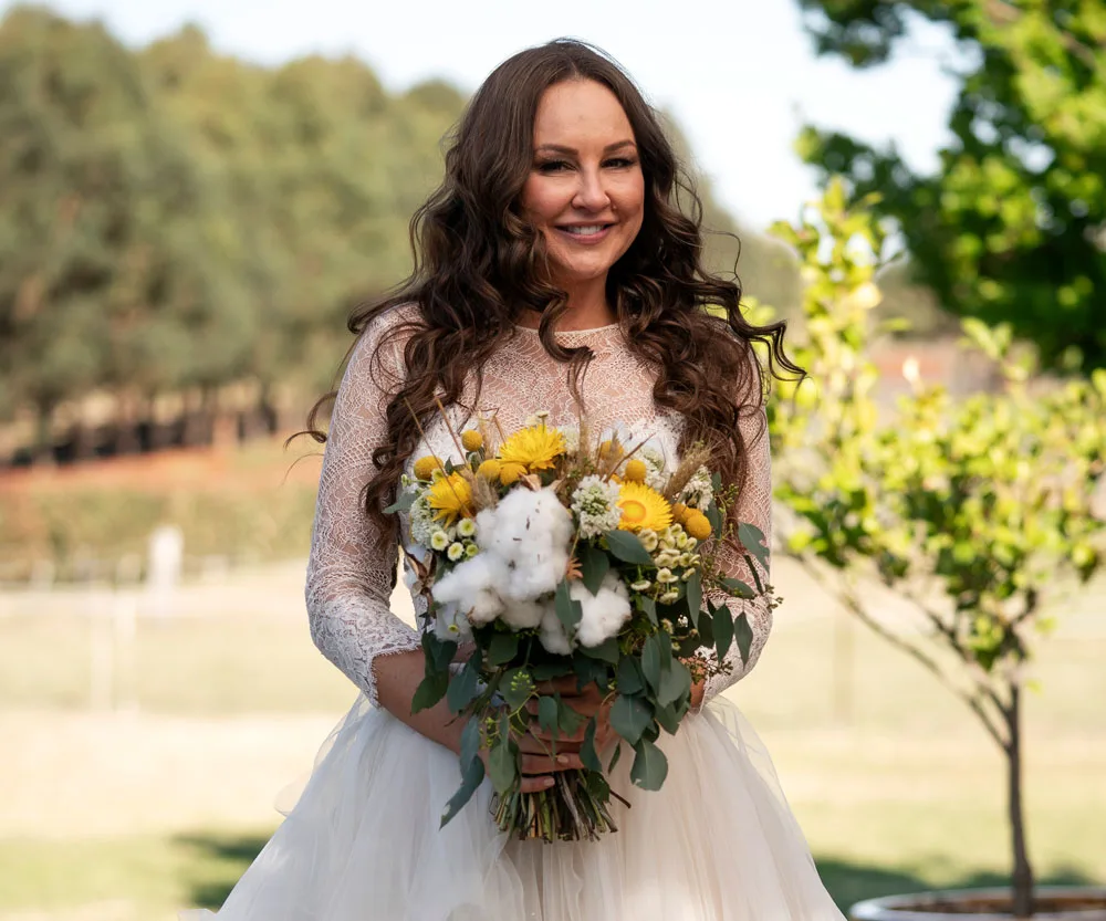 Bride in a lace dress holding a bouquet of sunflowers, smiling outdoors.