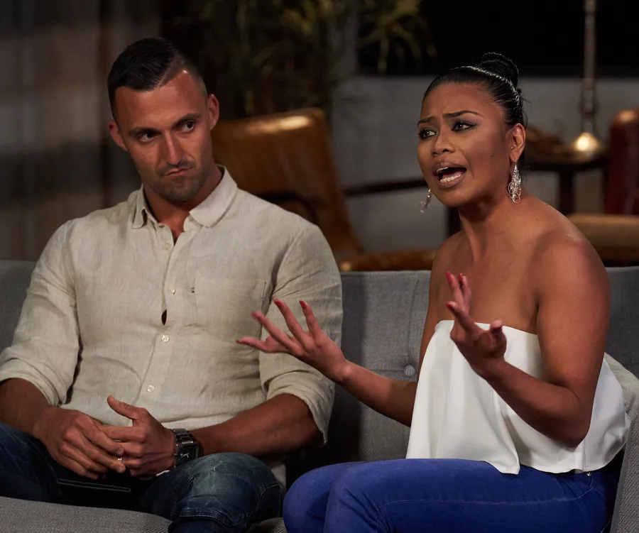 Man and woman sitting on a couch, woman gesturing angrily, man looking pensive, from a reality TV setting.