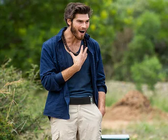 Man in blue shirt and beige pants, expressing surprise outdoors, likely reacting to an announcement or event.