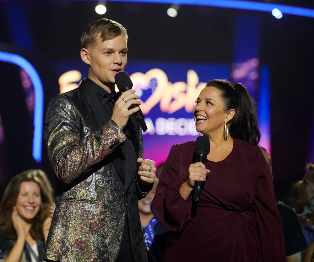 Two hosts with microphones on stage, smiling, during a live Eurovision event.
