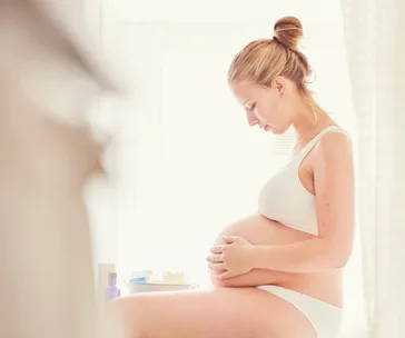 Pregnant woman sitting on a bed, wearing white undergarments, holding her belly and looking down pensively.