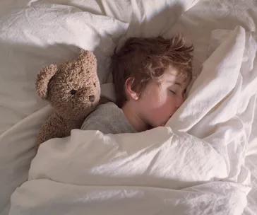 Child sleeping in bed with a teddy bear, wrapped in a white blanket.