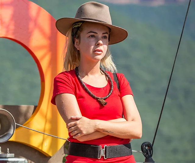 A woman in a red shirt and hat stands outdoors, arms crossed, with a serious expression.