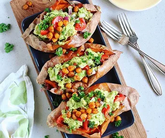 Baked sweet potatoes topped with mashed avocado, cherry tomatoes, chickpeas, and parsley on a black platter.