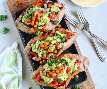 Baked sweet potatoes topped with mashed avocado, cherry tomatoes, chickpeas, and parsley on a black platter.