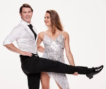 Two dancers in glamorous outfits posing happily for "Dancing with the Stars," with one lifting their leg playfully.