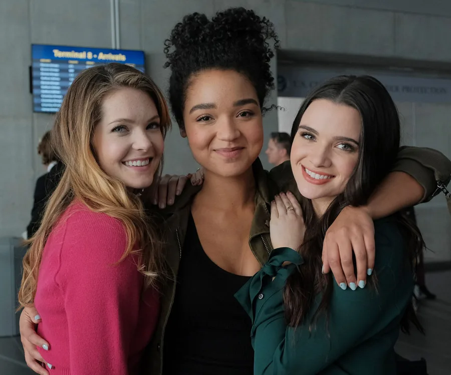 Three women smiling and hugging at an airport, with a terminal sign in the background.