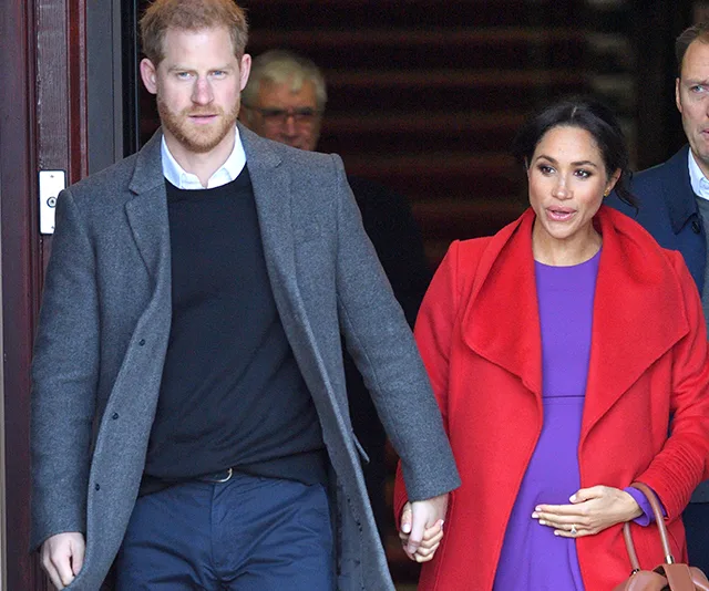 Royal couple holding hands, Meghan in purple dress and red coat, Harry in gray coat, exiting a building.