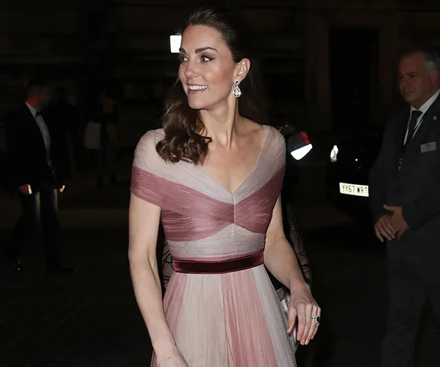 A woman in a pink Gucci dress with an off-shoulder design, smiling, and wearing drop earrings at an evening event.