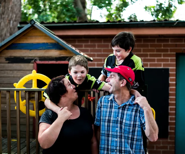 A family of four smiling and talking outside near a playhouse, with trees and a brick wall in the background.