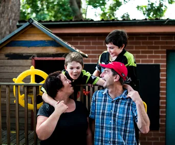 A family of four smiling and talking outside near a playhouse, with trees and a brick wall in the background.