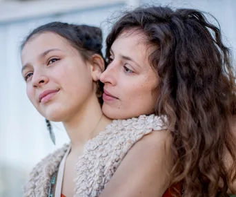 Mother embraces her teenage daughter lovingly, both with serene expressions and casual attire, outdoors.