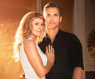 A man and a woman embrace in warm lighting, looking towards the camera. They are indoors with decorative elements in the background.