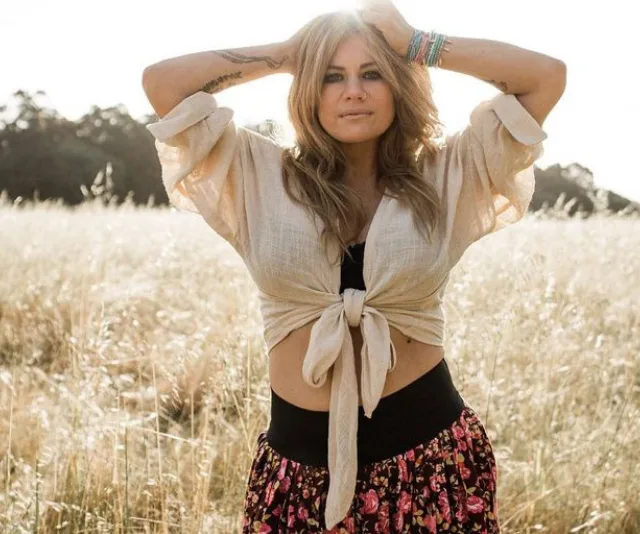 Woman in a field, wearing a tied shirt and floral skirt, with hands on head, looking at camera.