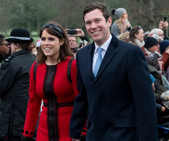 A smiling couple, with the woman in red and the man in a dark coat, walking outdoors surrounded by a crowd.