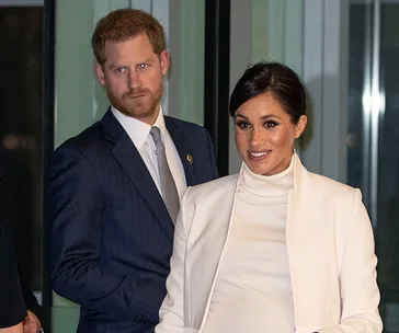 Two individuals, one wearing a dark suit and the other in a white outfit, standing indoors, smiling at the camera.