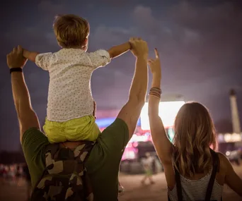 A family at an outdoor music festival at dusk, child on shoulders, facing a lit-up stage.