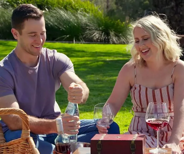 Couple enjoying a picnic on grass, man opening water bottle, woman holding wine glass, surrounded by picnic items.