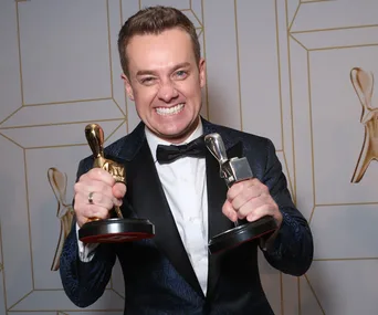 Smiling person in a tuxedo holding two Logie awards, celebrating victory at the 2019 TV Week Logie Awards.