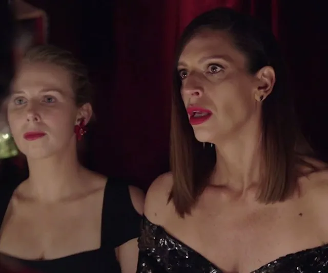 Two women in evening attire look intently off-camera, in a dimly lit setting.