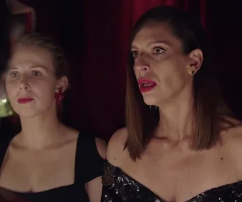 Two women in evening attire look intently off-camera, in a dimly lit setting.