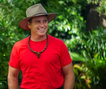 Man in a red shirt and hat smiles outdoors in a jungle setting.
