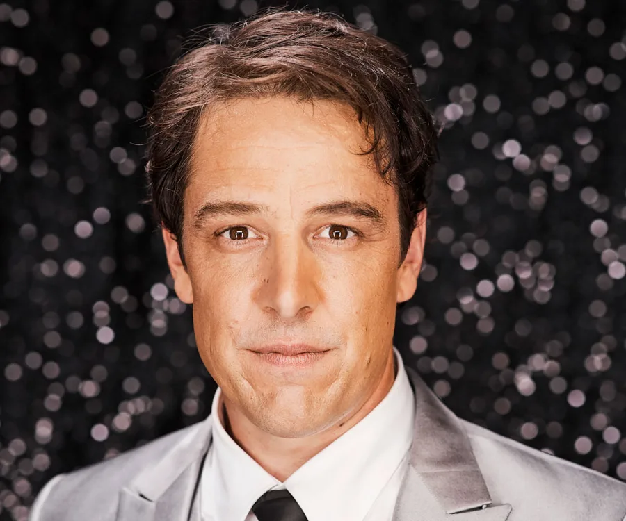 "Man in a shiny suit with a speckled background, looking directly at the camera."