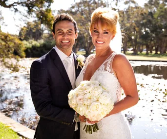 Bride and groom smiling outdoors by a pond, bride holding a bouquet of white roses.