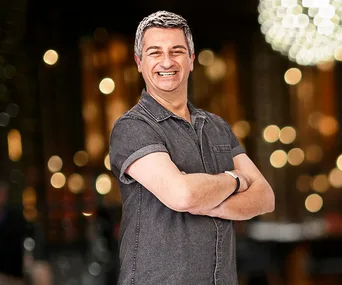 Smiling man with crossed arms in casual denim shirt, blurred lights in background.