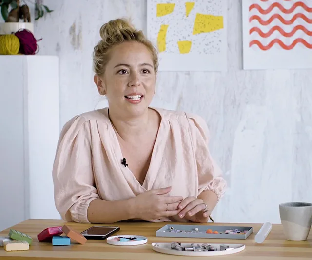 A woman sits at a craft table with tools and colorful materials, smiling, against a light background with abstract wall art.