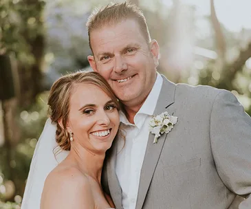 Bride and groom smiling together outdoors on their wedding day, bride in white and groom in a grey suit.