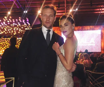 A couple dressed elegantly pose at a glamorous event with bright, decorative lighting in the background.