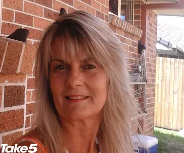 A woman with long blonde hair smiling, standing outside near a brick wall.