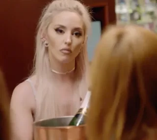 A woman with long blonde hair holds an ice bucket, looking serious during a conversation indoors.