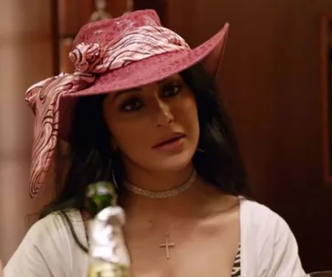 Woman wearing a pink patterned hat with a bow and cross necklace, sitting in a room with dim lighting.