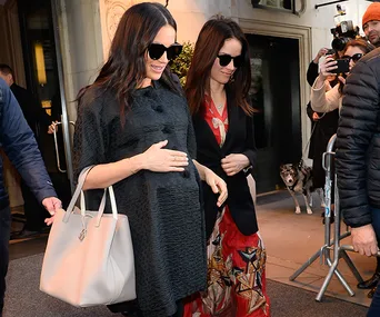Pregnant woman in black coat, sunglasses, and holding a bag, walking with another woman outside a building.