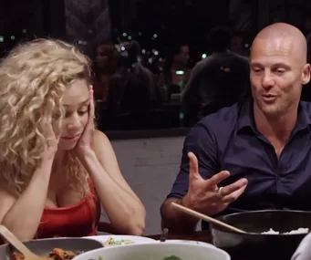 A woman with curly hair looks distressed while a bald man gestures during a dinner at "Married at First Sight."