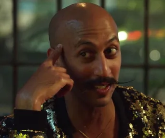 A person with a mustache wearing a sequined jacket, gesturing with a finger to their temple, smiling.