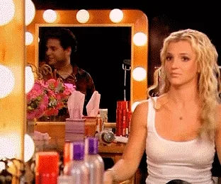 A person sitting at a makeup table with lighted mirror, surrounded by beauty products.